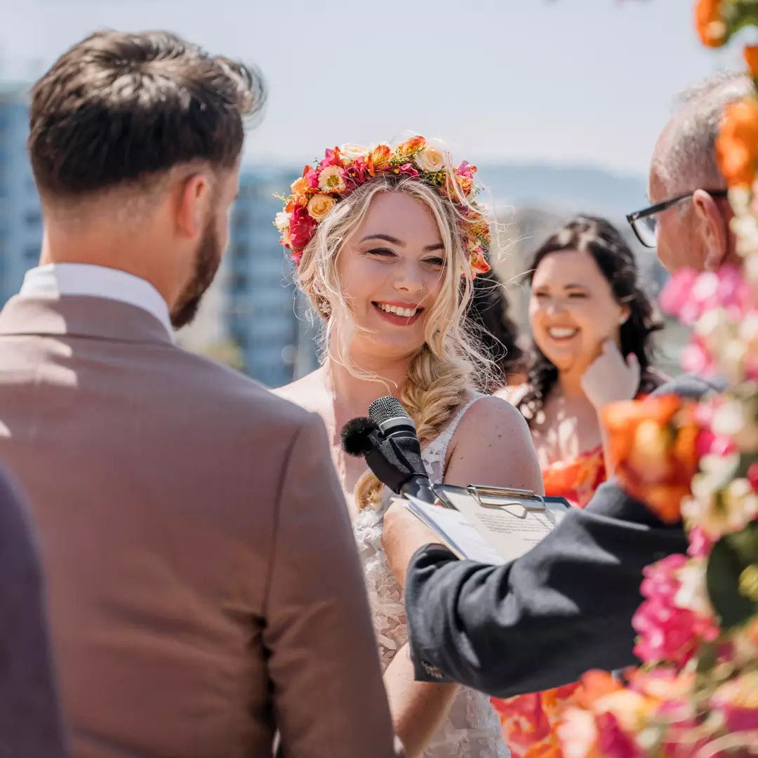 A bride with a floral crown smiles during an outdoor wedding ceremony, facing the groom, with an officiant holding a microphone and clipboard nearby.