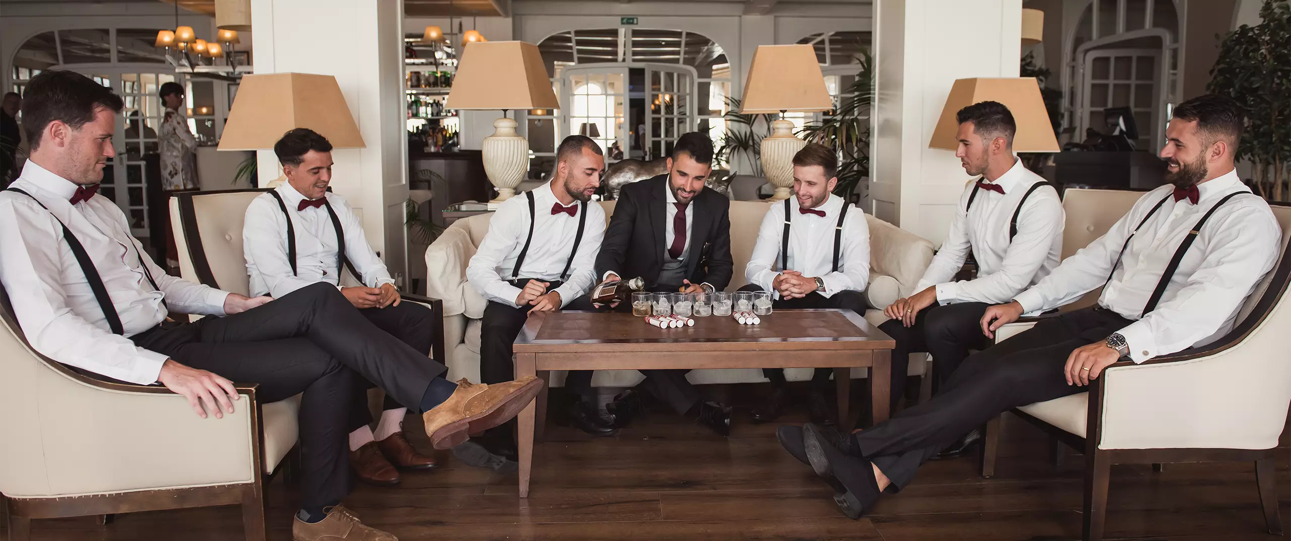 Six men in dress shirts, bow ties, and suspenders sit around a coffee table in a lounge, appearing to play a board or card game, with glasses and chips on the table.