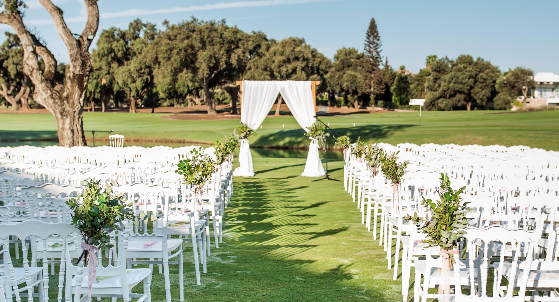 Rows of white chairs arranged on grass face a draped wedding arch, with greenery decorations and trees in the background on a sunny day.