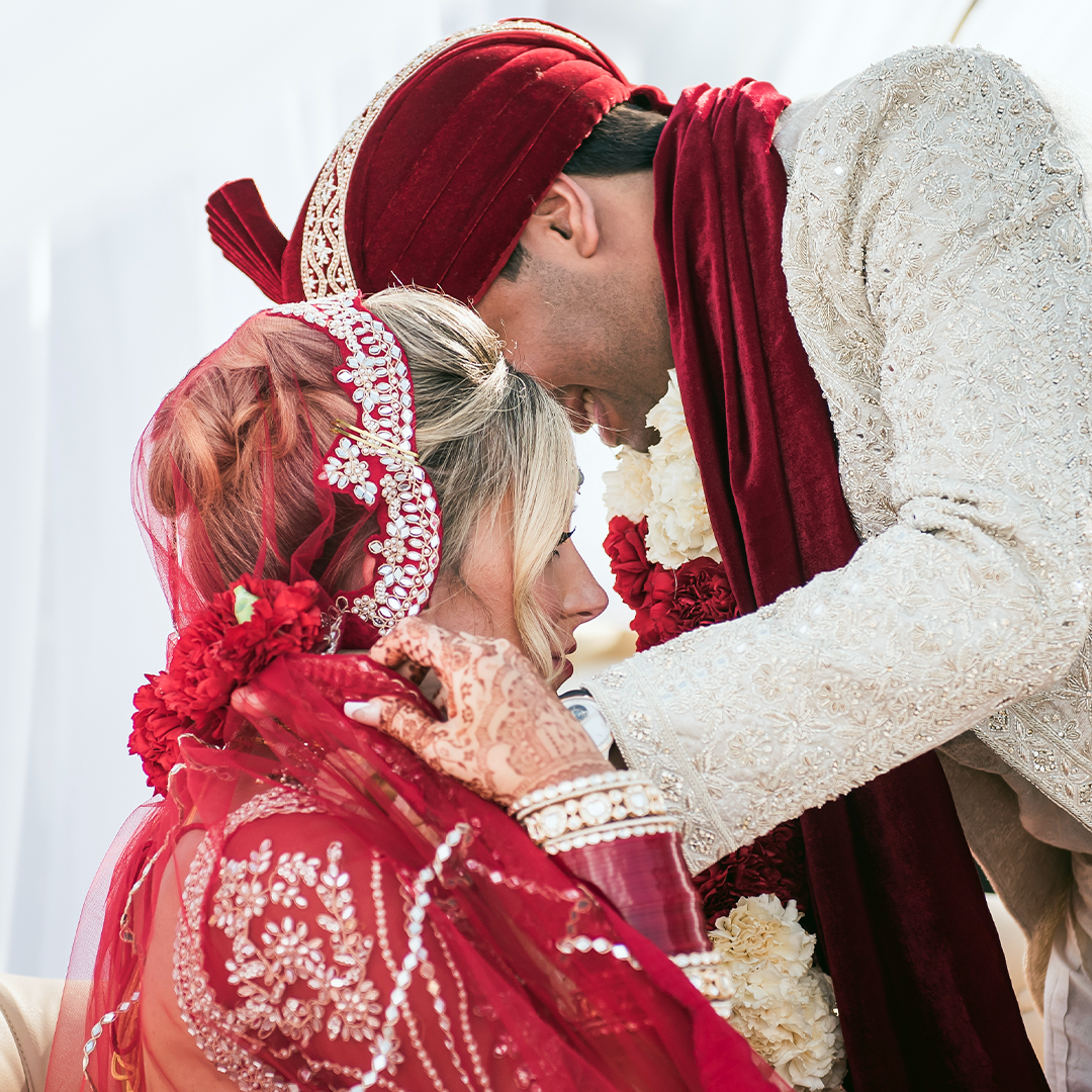 A bride and groom in traditional South Asian wedding attire participate in a ceremony, with the groom adjusting the bride's veil.