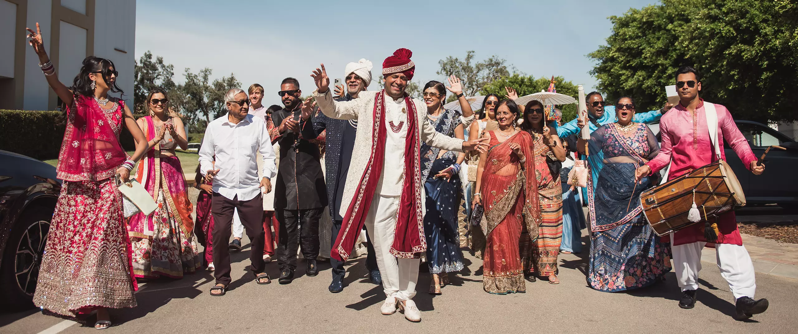A groom in traditional Indian attire leads a joyful wedding procession outdoors, surrounded by family and friends in colorful clothing.