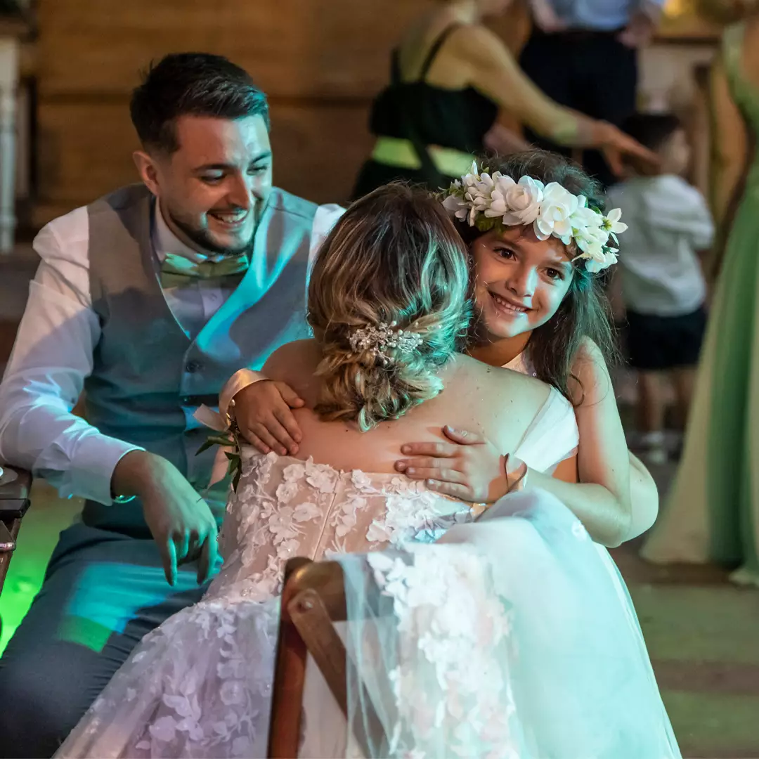 A bride hugs a young girl wearing a flower crown while a man in a vest and bowtie smiles beside them at a wedding reception.