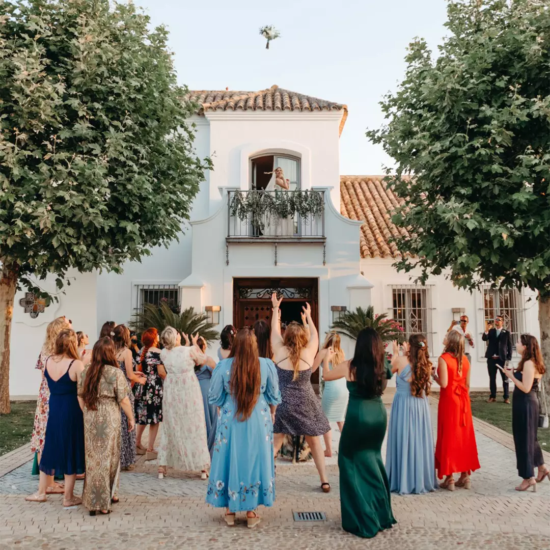 A group of women stand outside a white house, reaching up as a bride on a balcony throws her bouquet.