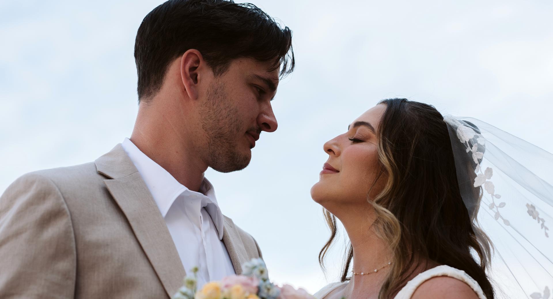 A bride and groom stand close together outdoors, facing each other. The bride wears a veil and holds a bouquet; the groom wears a beige suit.