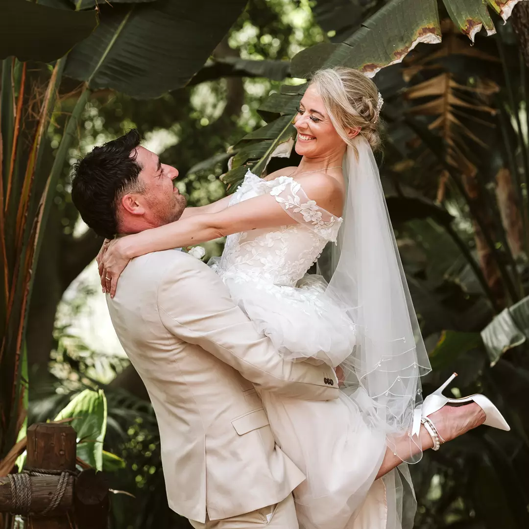A groom in a light suit lifts a smiling bride in a white wedding dress and veil, surrounded by lush green foliage outdoors.