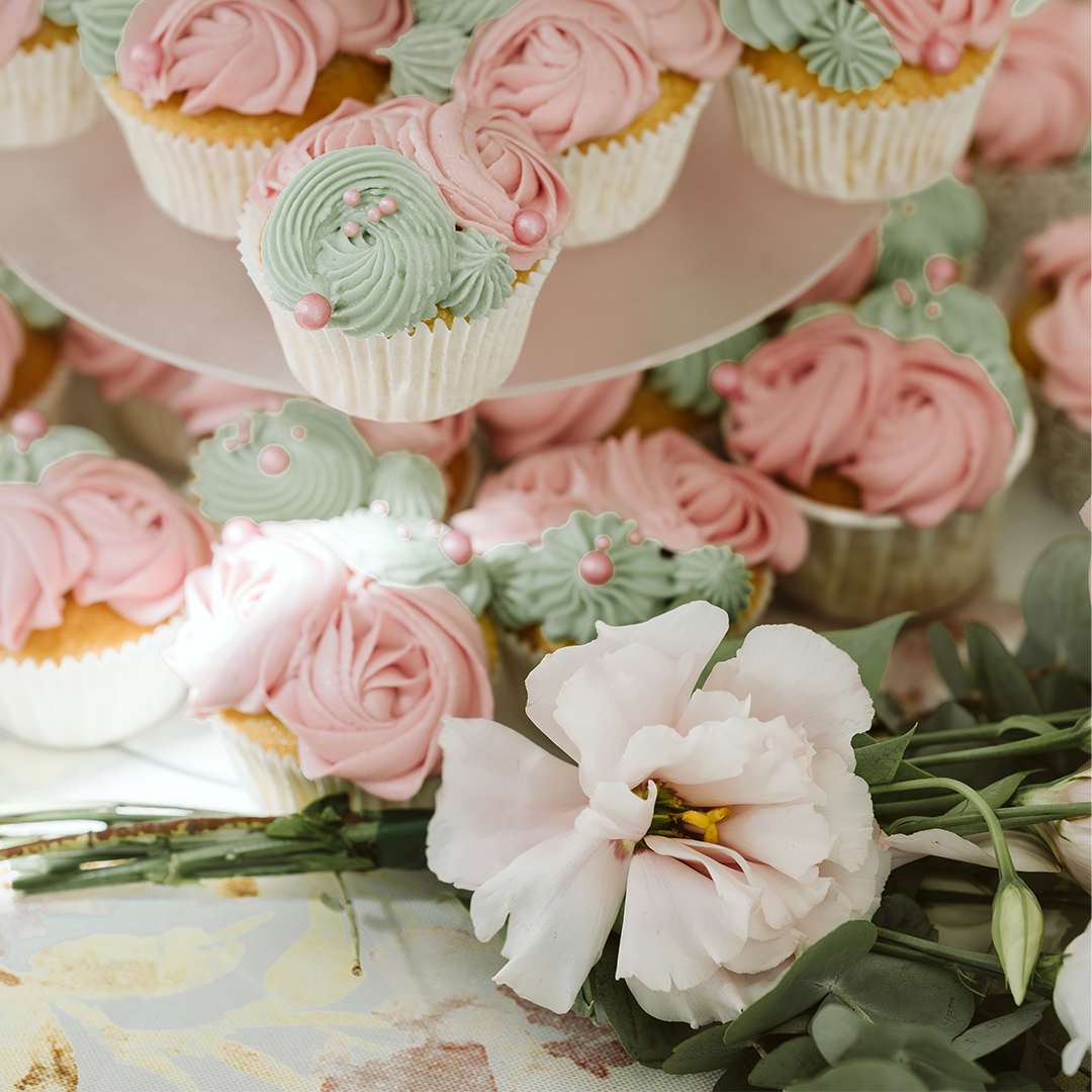 Cupcakes decorated with pink and green frosting arranged on a tiered stand, with a white flower and greenery placed beside them on a table.