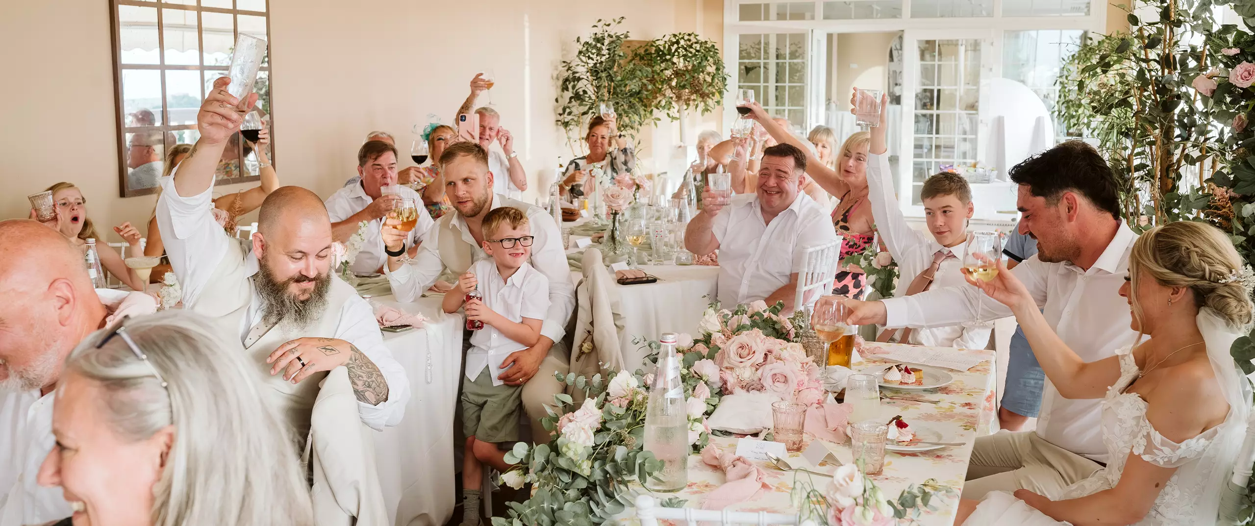 A group of people dressed in white raise glasses in a toast at a decorated wedding reception with flowers and table settings.
