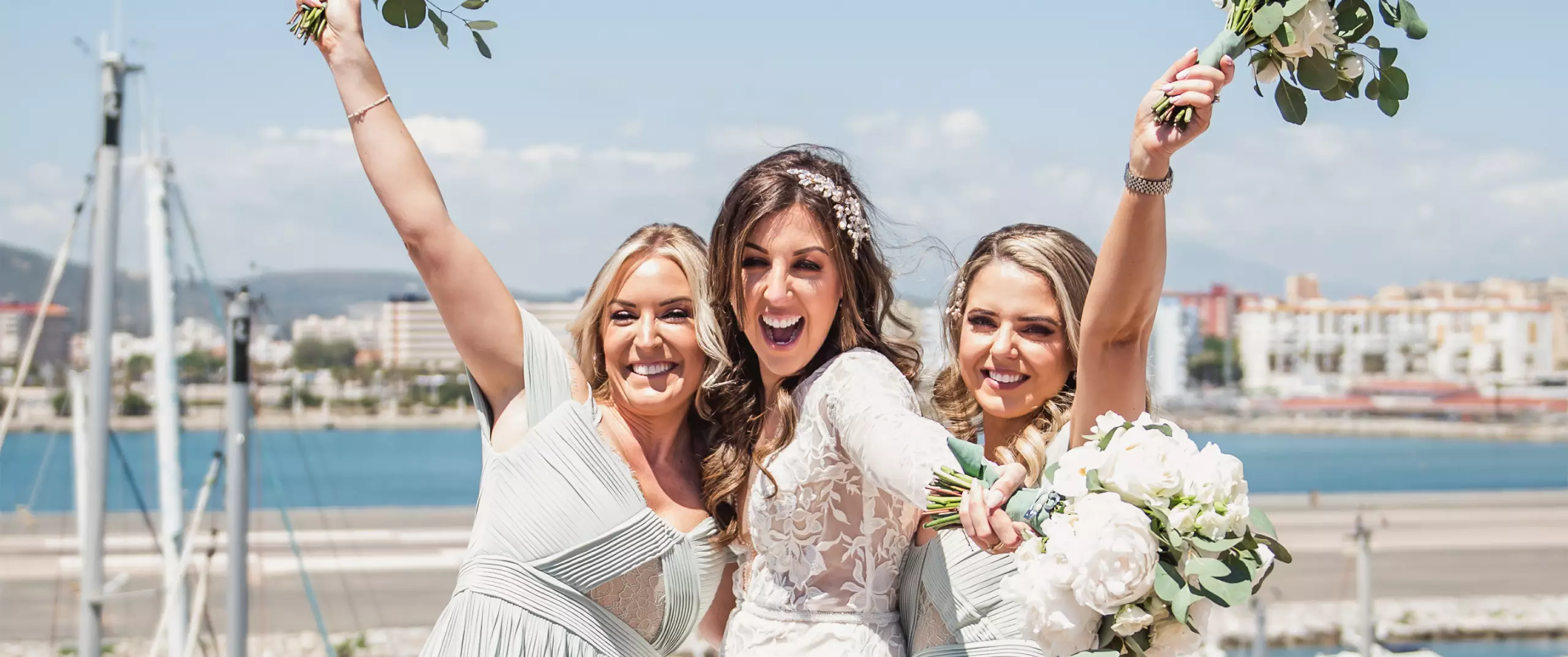 Three women in light-colored dresses, including a bride in white holding a bouquet, pose outdoors with arms raised, smiling in front of a waterfront cityscape.
