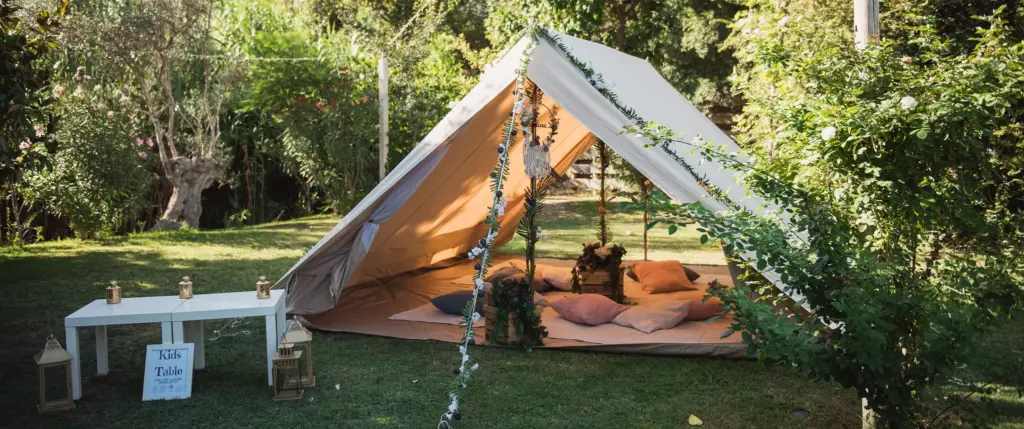 A beige canvas tent with cushions inside is set up on grass, surrounded by greenery. A low white table with lanterns sits nearby.