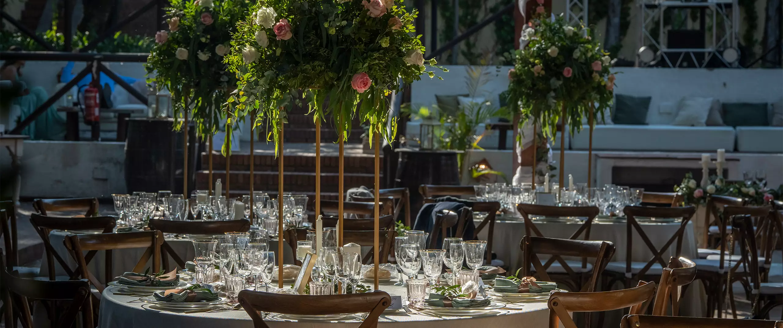 Round tables are set for an event with glassware, plates, and napkins, each centerpiece featuring tall arrangements of greenery and flowers. Wooden chairs surround the tables.