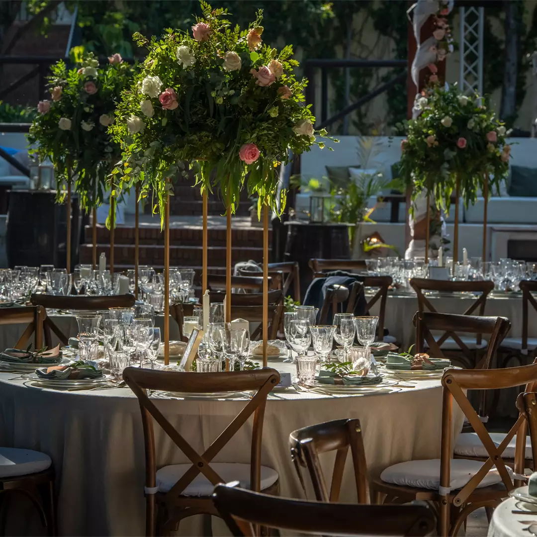 A round table set for an outdoor event with glassware, plates, and a tall floral centerpiece, surrounded by wooden chairs.
