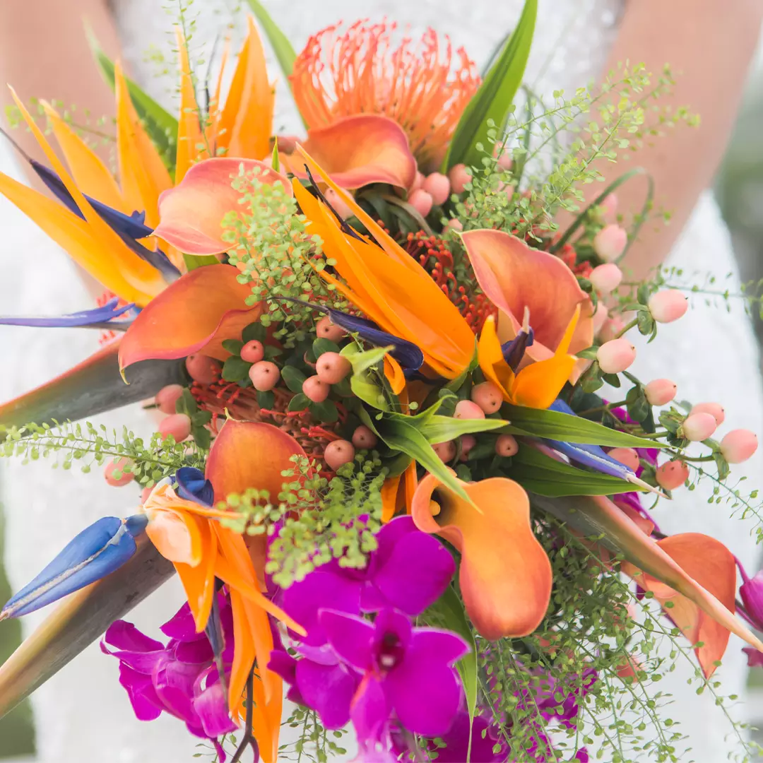 A person in white holds a vibrant bouquet featuring orange calla lilies, birds of paradise, purple orchids, pink berries, and greenery.