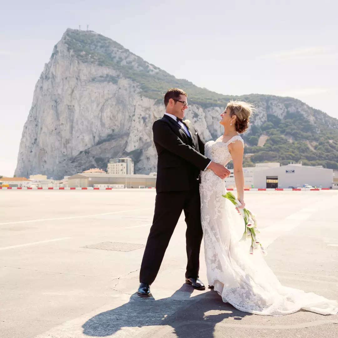 A bride and groom in formal attire stand facing each other, smiling, with the Rock of Gibraltar visible in the background.