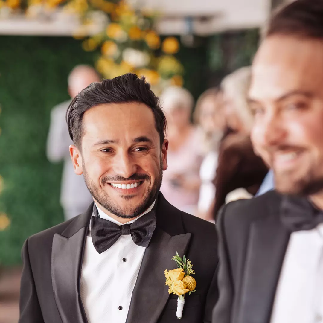 Two men in tuxedos stand together at an event. One man is smiling at the other. Both wear black bow ties, and one has a yellow boutonniere. People and greenery appear in the background.