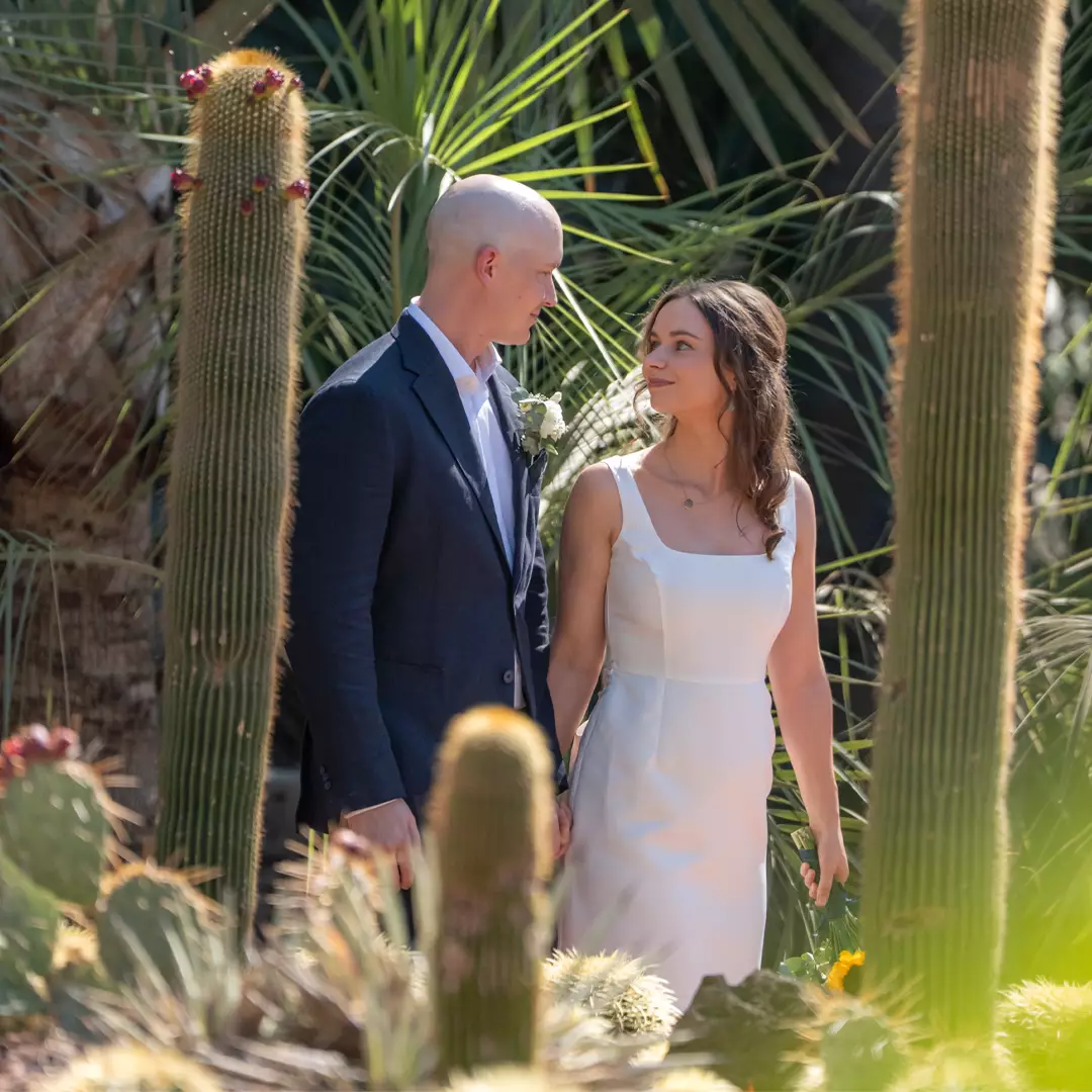 A couple dressed in formal attire stands together among tall cacti and desert plants, looking at each other and holding hands.