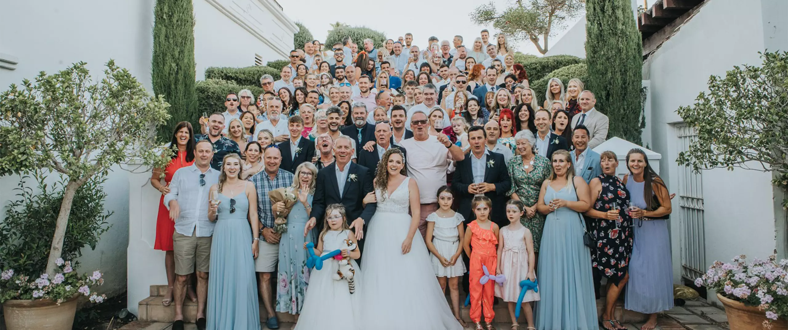 A large wedding group photo with the bride and groom in the center, surrounded by guests of various ages, standing on outdoor steps.