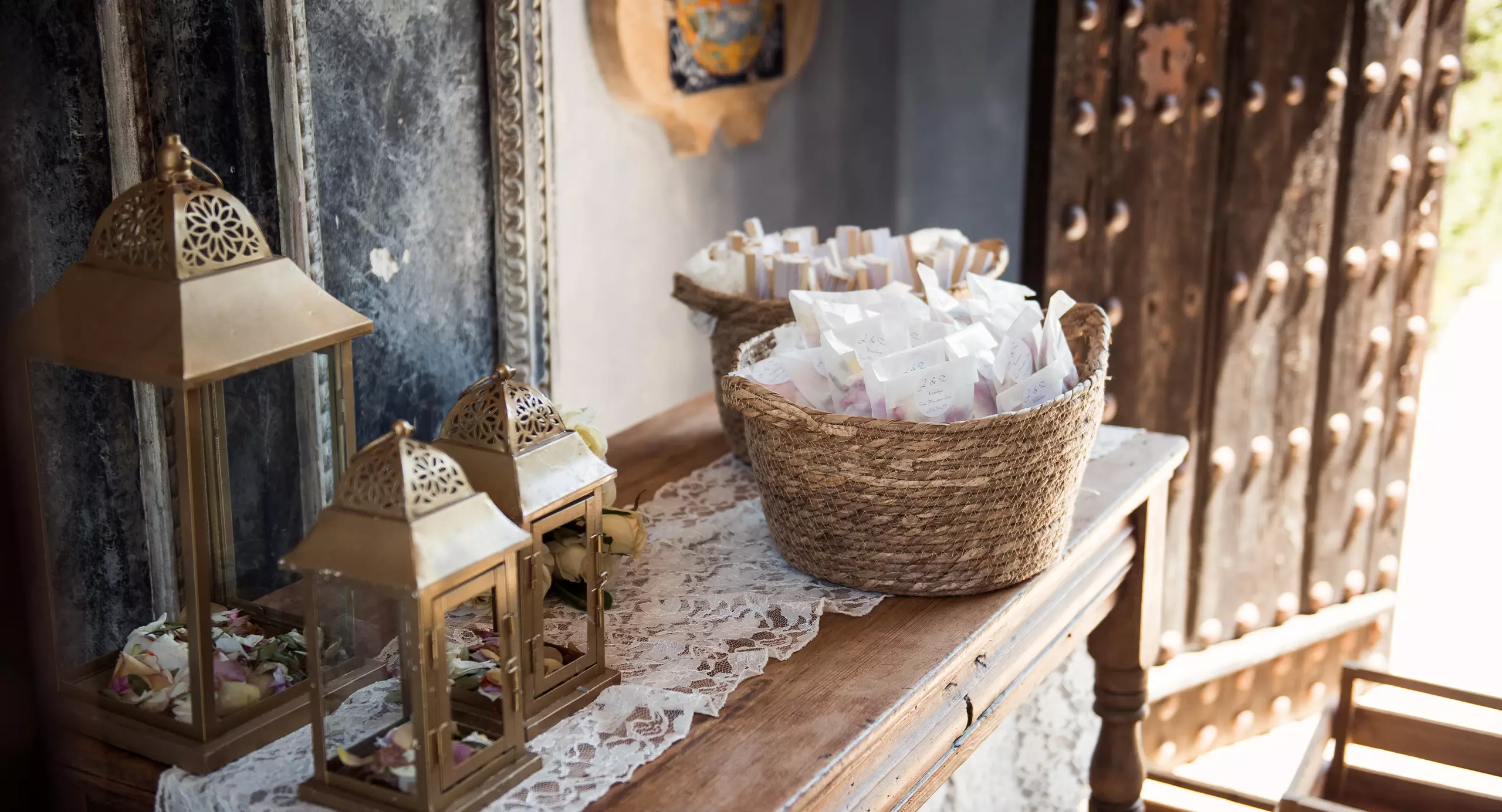 A woven basket filled with small paper packets sits on a lace-covered wooden table next to two brass lanterns in a rustic setting.