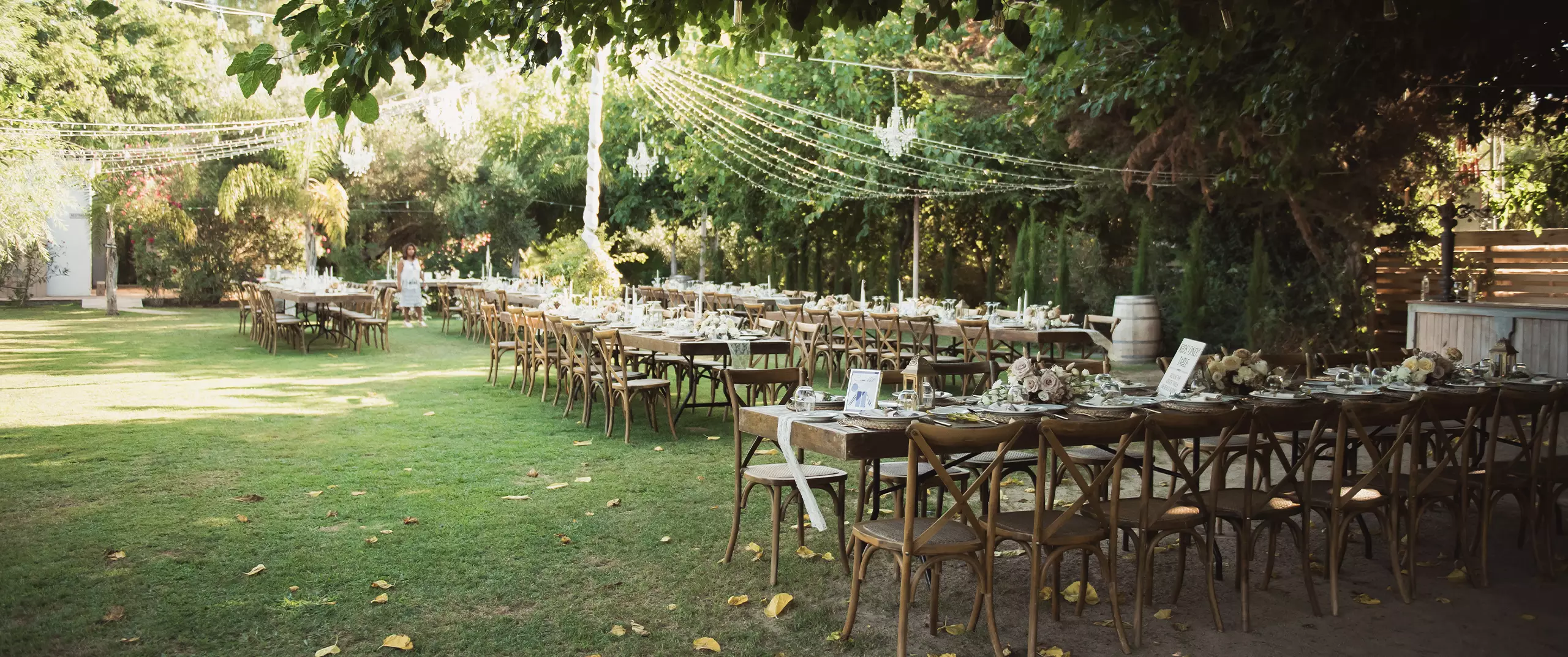 Long wooden tables and chairs are set up outdoors on a lawn under string lights, ready for an event, with greenery and trees surrounding the area.
