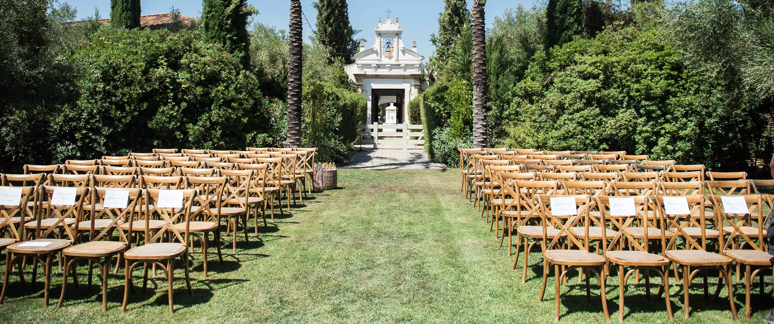Rows of wooden chairs are arranged on a grassy lawn facing a small white building, set up for an outdoor event or ceremony, with trees and bushes surrounding the area.