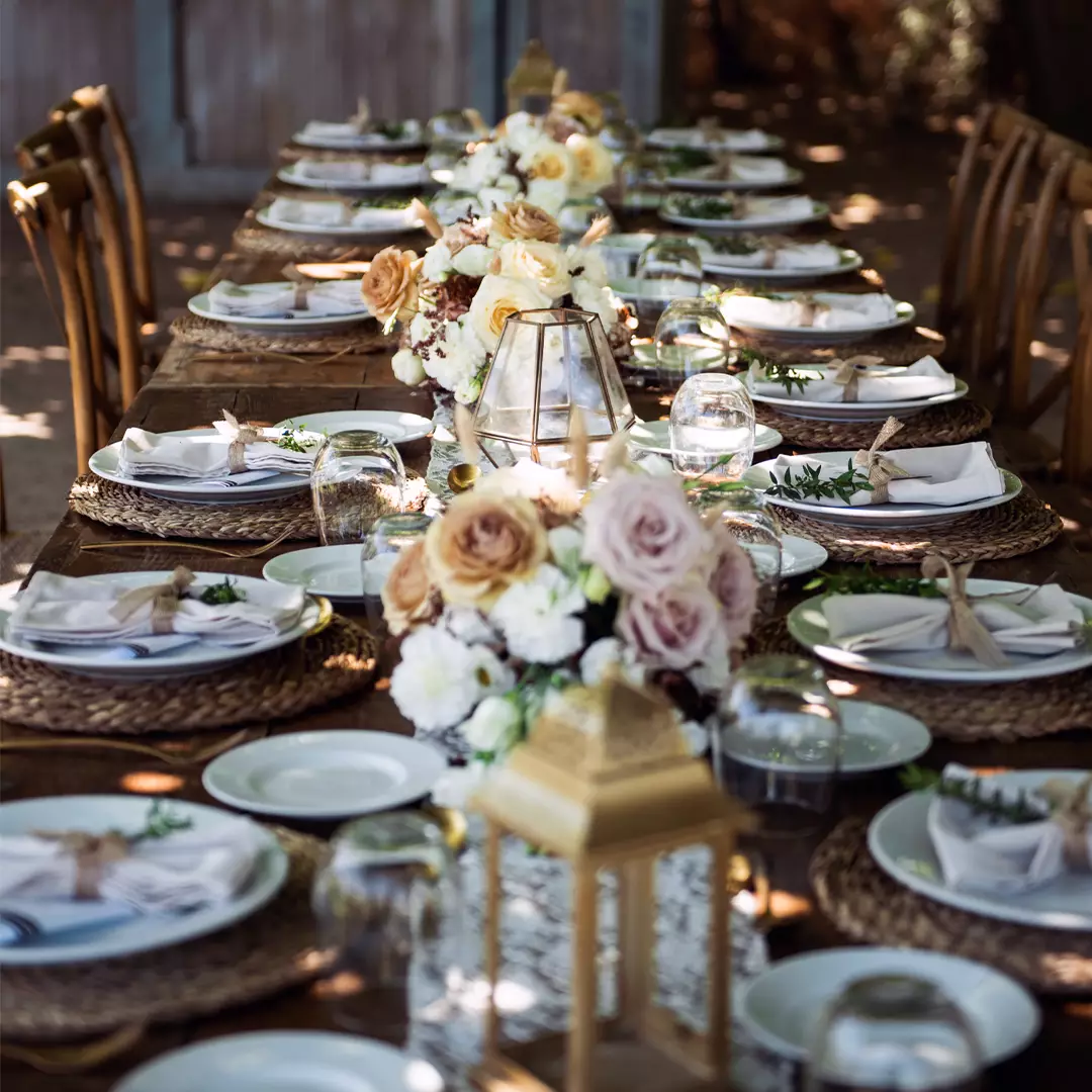 A long, outdoor dining table is set with white plates, woven placemats, folded napkins, glassware, and floral centerpieces in soft, neutral tones.