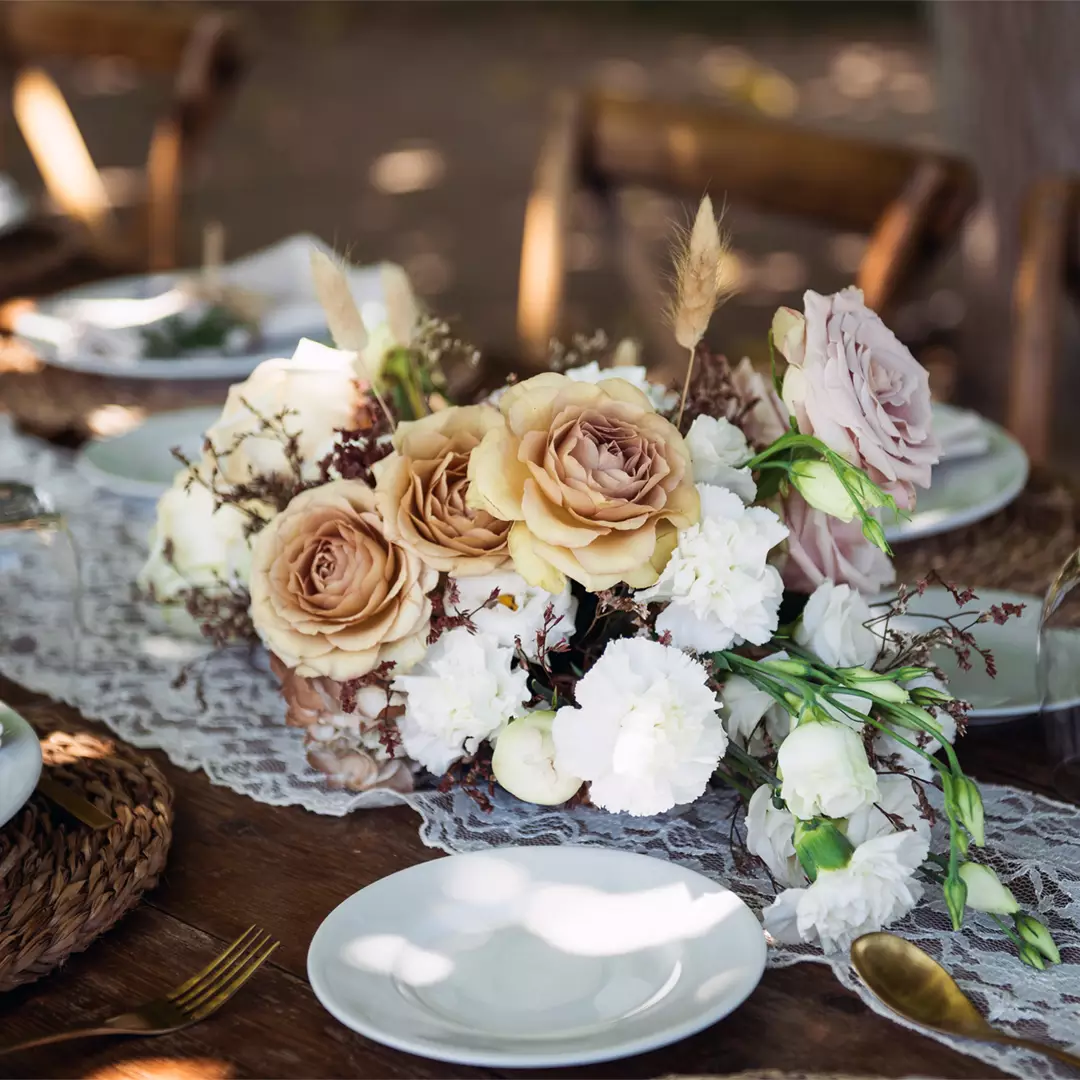 A floral centerpiece with beige and white roses, greenery, and lace accents sits on a wooden table set with white plates and woven placemats.