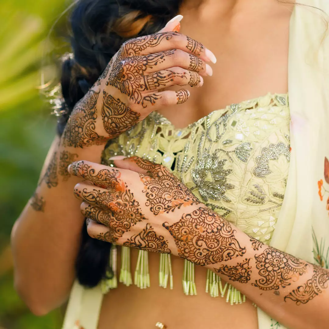 A woman wearing a light green, embroidered outfit displays intricate henna designs on both hands and arms, with a blurred outdoor background.