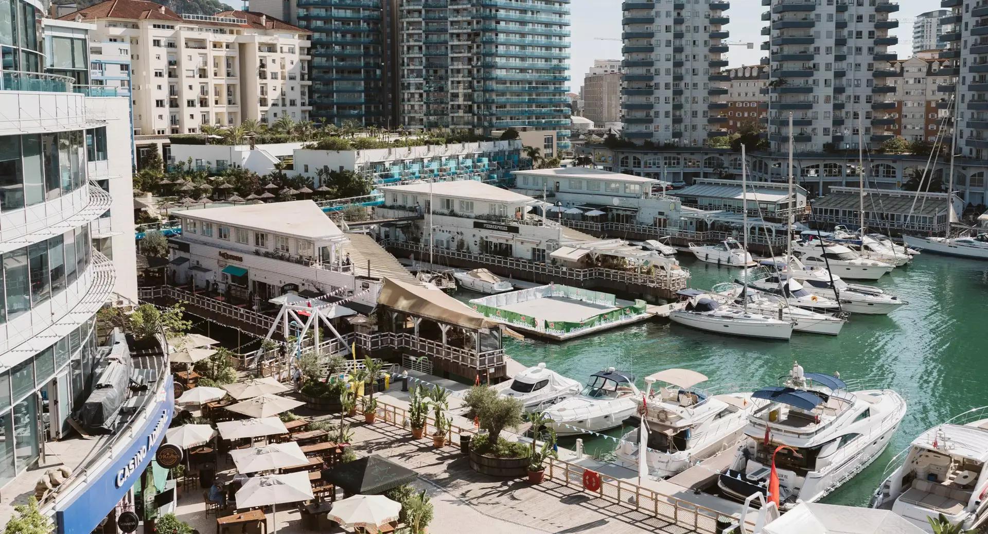 A marina with several docked boats, waterfront restaurants with outdoor seating, and modern high-rise buildings in the background under daylight.