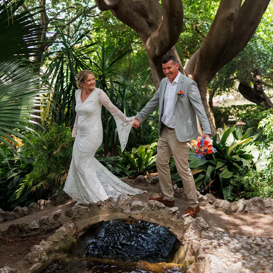 A bride and groom hold hands and smile while walking over a small stone bridge in a lush garden setting.