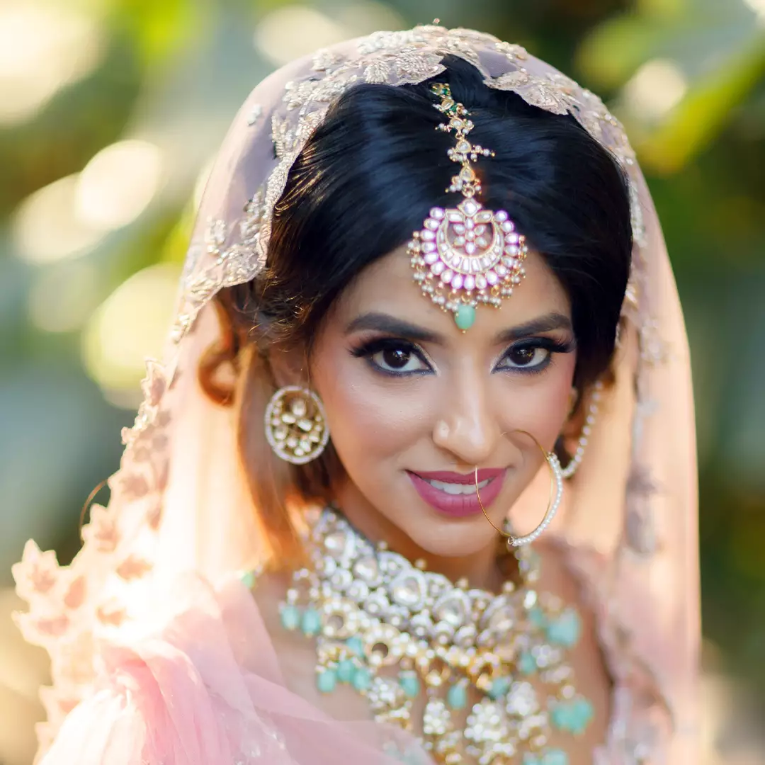 A woman wearing traditional bridal attire with intricate jewelry, including a maang tikka, earrings, necklace, and a sheer embroidered veil, smiling at the camera.