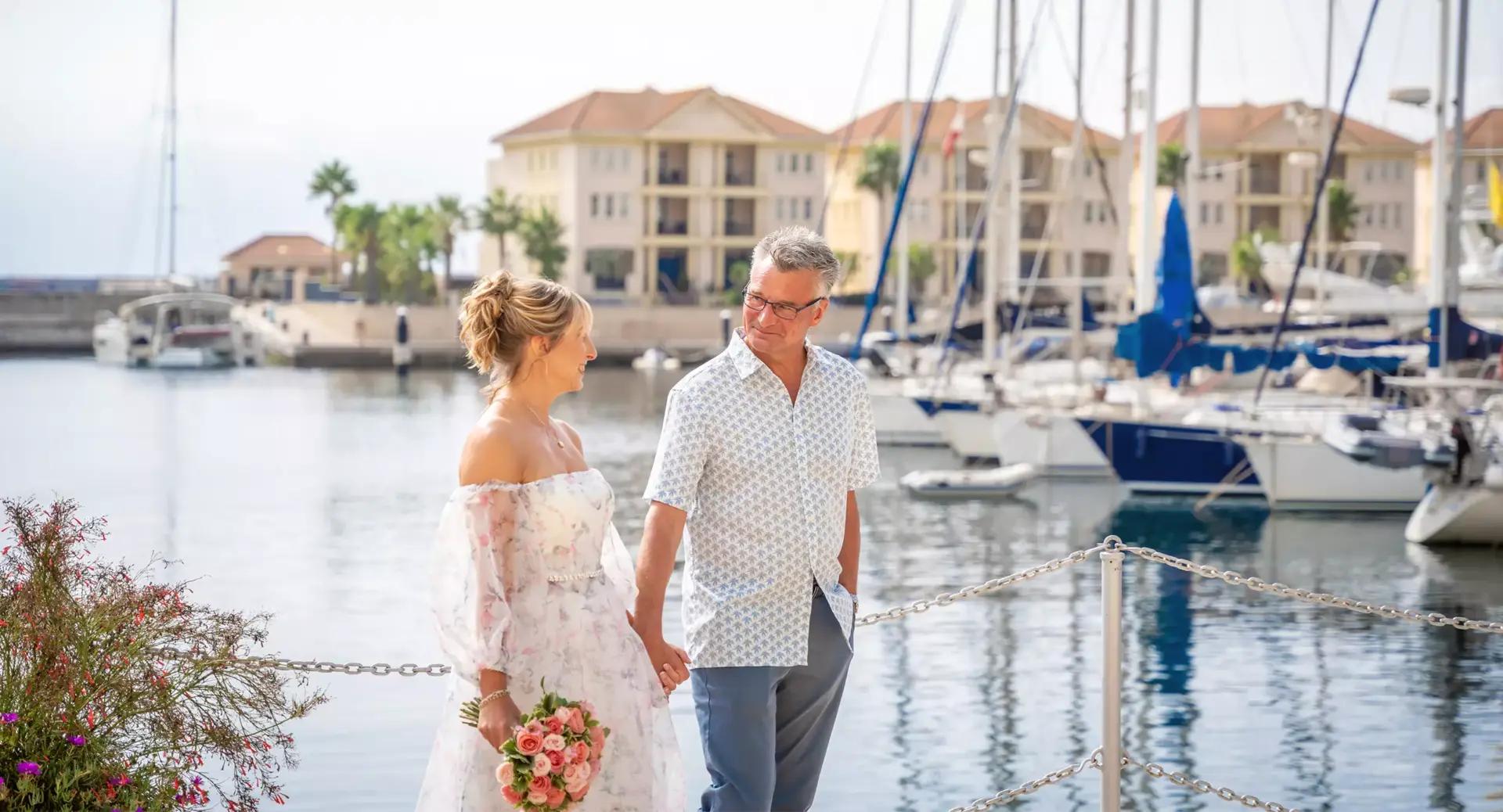 A couple holding hands and smiling at each other walks along a marina with sailboats and waterfront buildings in the background. The woman holds a bouquet of flowers.