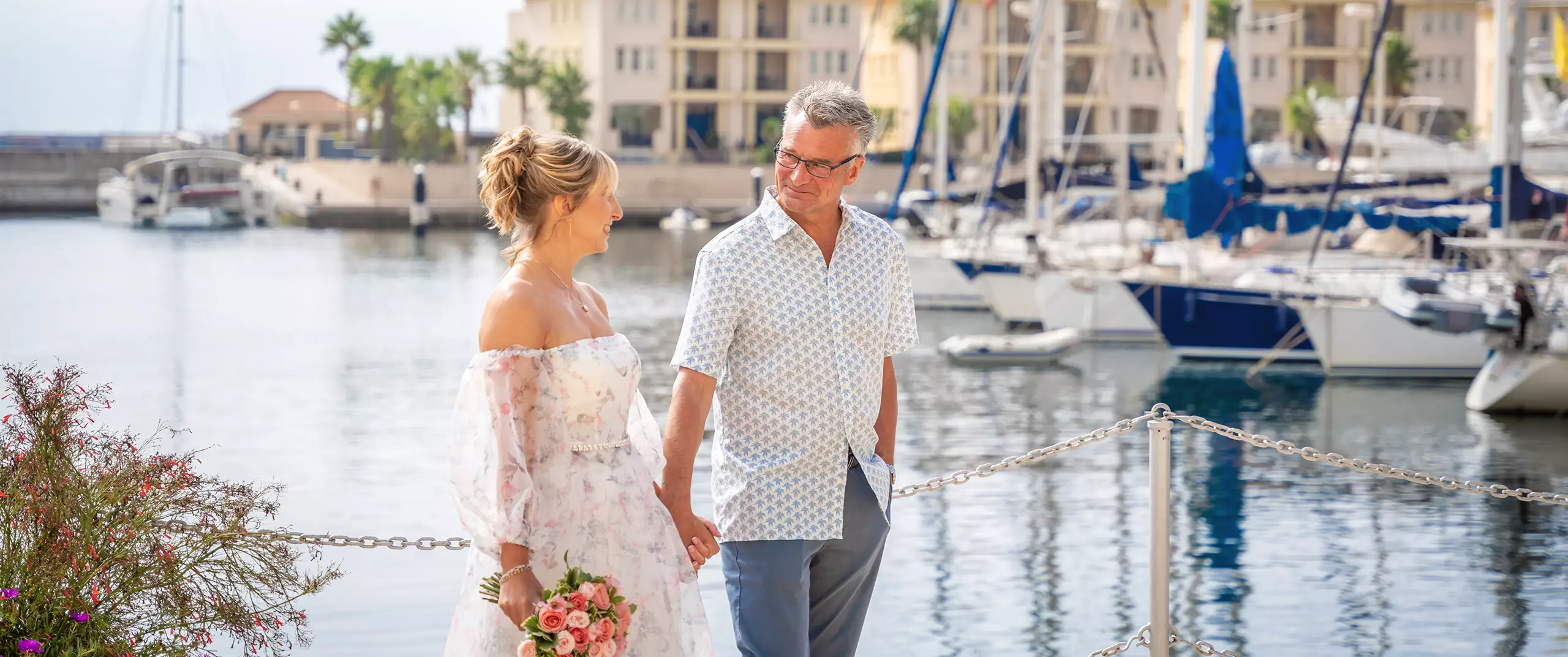 A couple holding hands and smiling at each other while walking by a marina, with yachts and buildings in the background. The woman holds a bouquet of flowers.