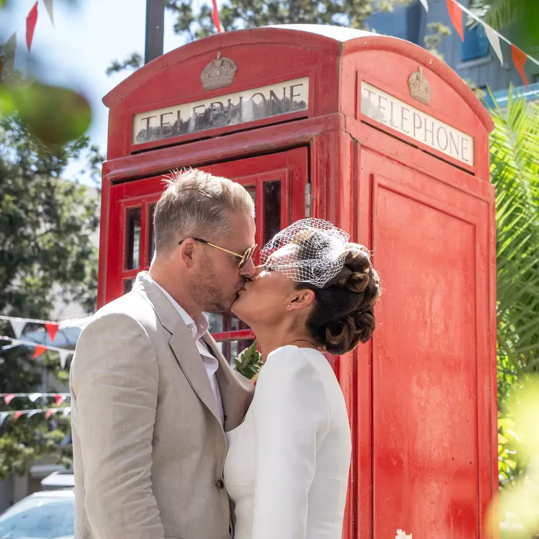 A newlywed couple kisses in front of a classic red British telephone booth decorated with bunting on a sunny day.
