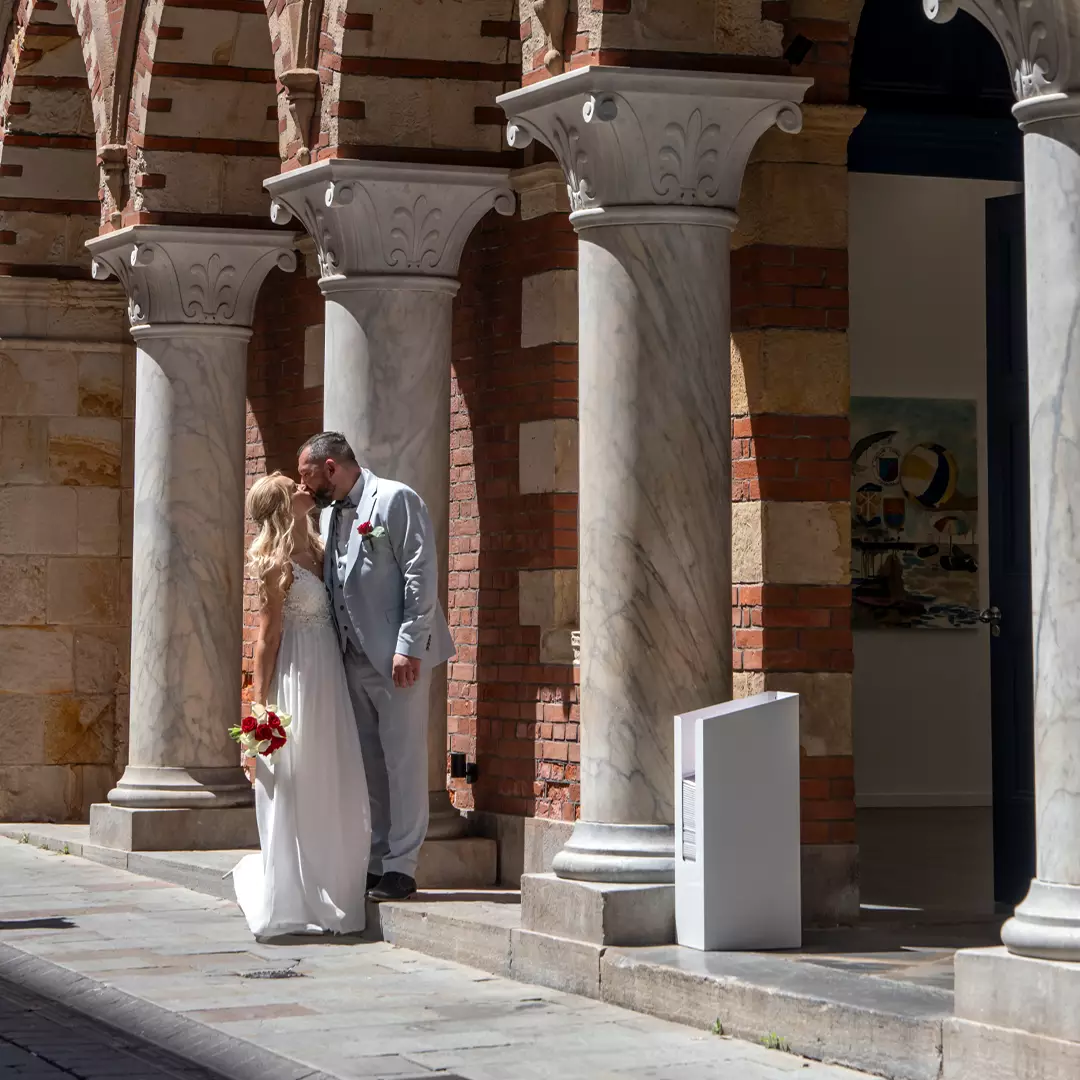 A bride and groom stand closely together, holding hands and flowers, under marble columns in front of a brick building.