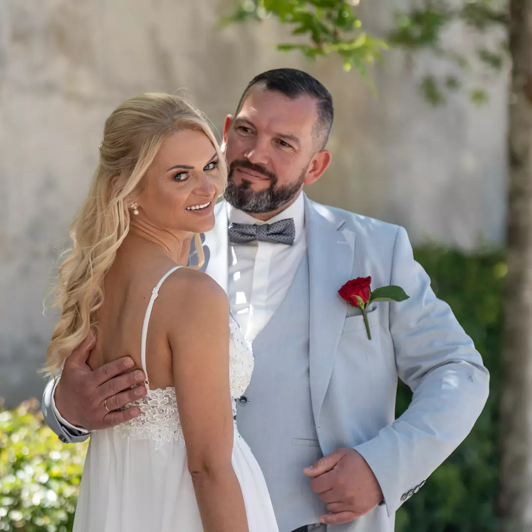 A bride in a white dress and a groom in a light suit with a red rose boutonniere pose together outdoors, smiling.