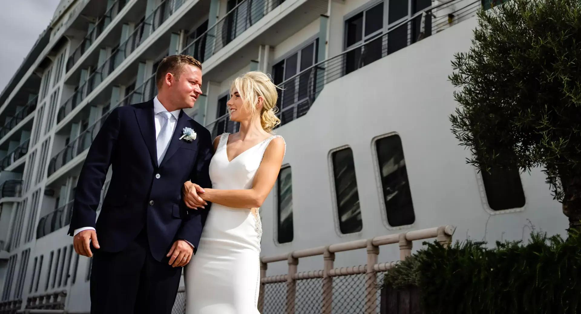A bride and groom in formal wedding attire stand arm in arm outside a large cruise ship, looking at each other and smiling.