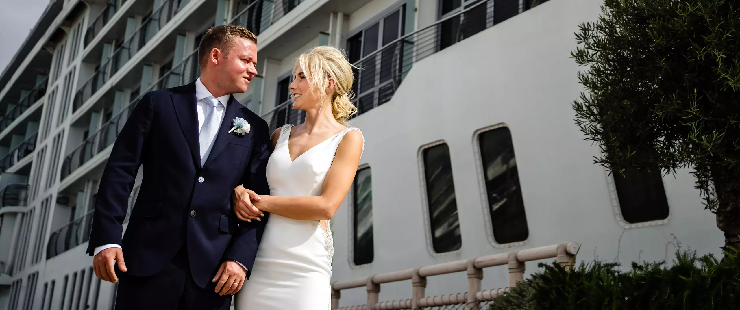 A bride and groom in formal attire stand arm in arm, smiling at each other, with a large white cruise ship in the background.