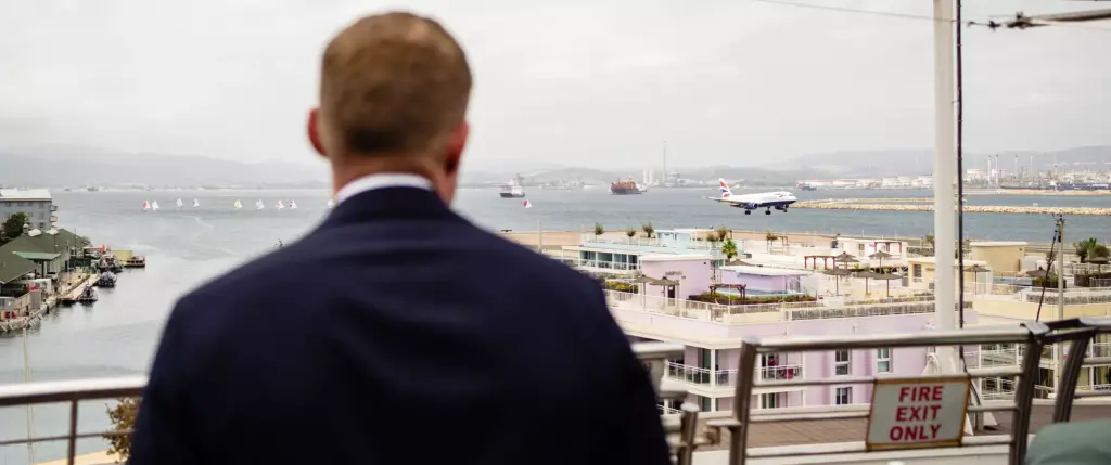 A man in a suit stands on a balcony overlooking a bay, with an airplane landing in the background and boats on the water.