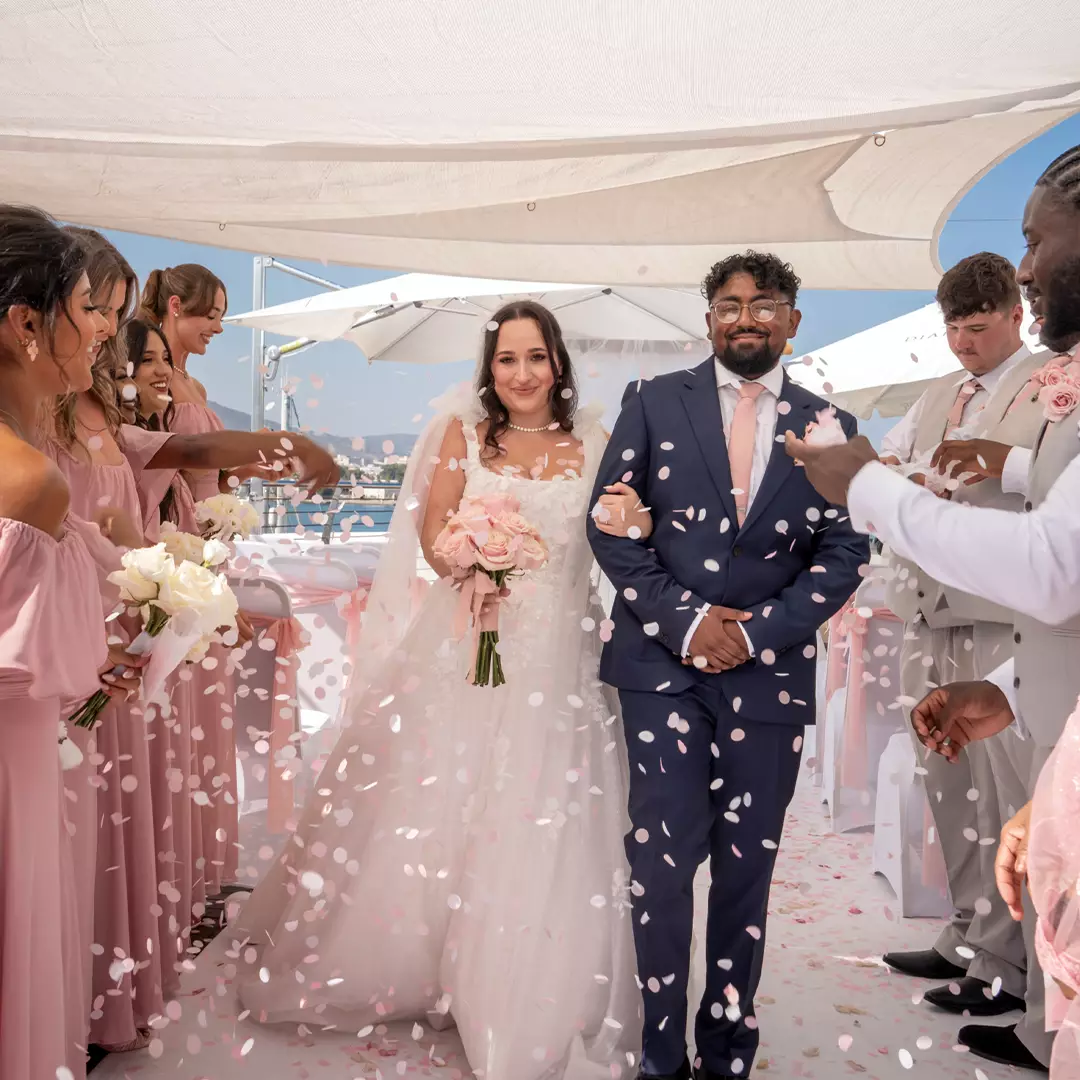A bride and groom walk down the aisle as guests in pink attire throw flower petals at an outdoor wedding ceremony under white canopies.