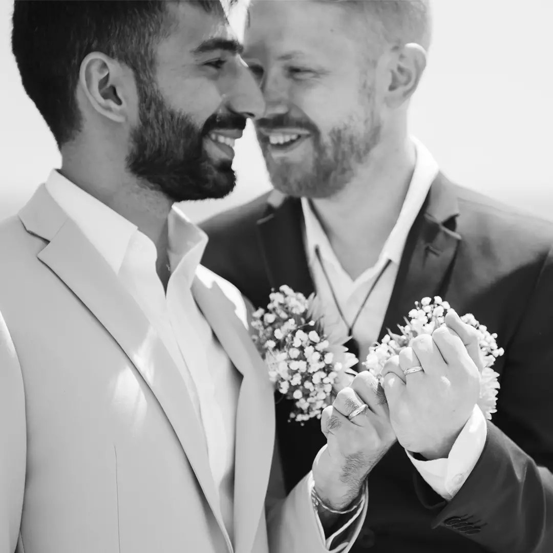 Two men in suits smile and hold hands with intertwined fingers, each holding a small bouquet of flowers, in a black and white photo.