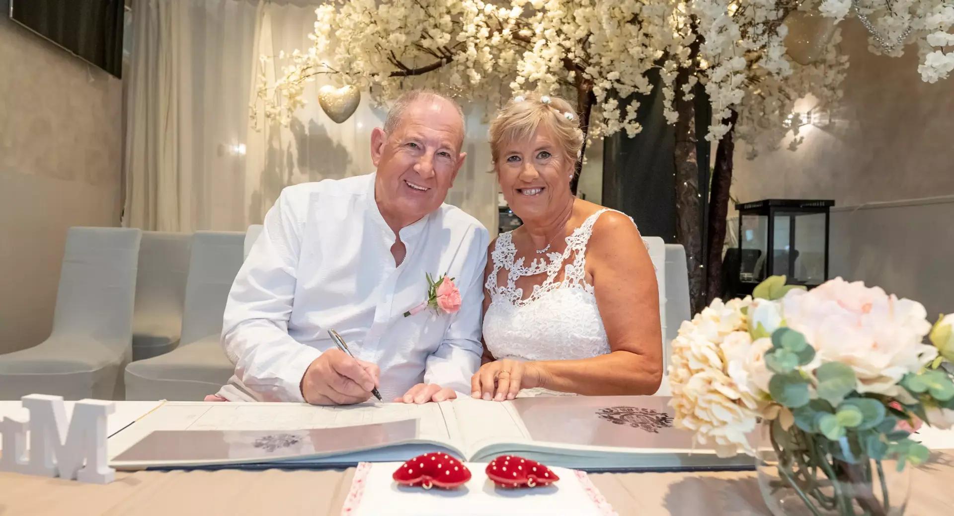 An older couple in wedding attire sits at a table, smiling as the man signs a document. White flowers and a bouquet decorate the scene.