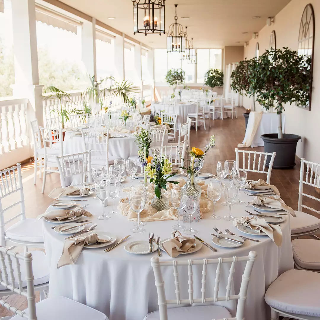 Elegant outdoor banquet setup with round tables covered in white linens, set with glassware and plates, and decorated with floral centerpieces and white chairs.