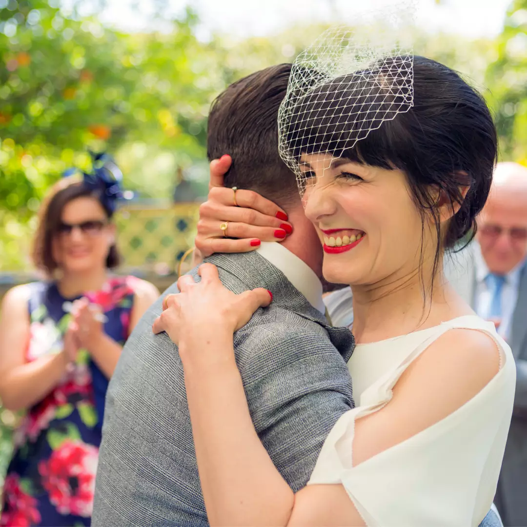 A smiling bride in a white dress hugs a man in a suit outdoors; guests in colorful attire applaud in the background.