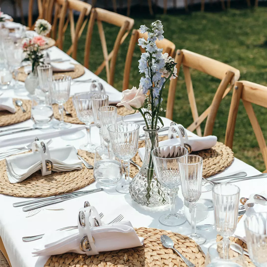 A long outdoor table is set with woven placemats, glassware, white napkins in rings, and vases of flowers on a white tablecloth. Wooden chairs line both sides of the table.