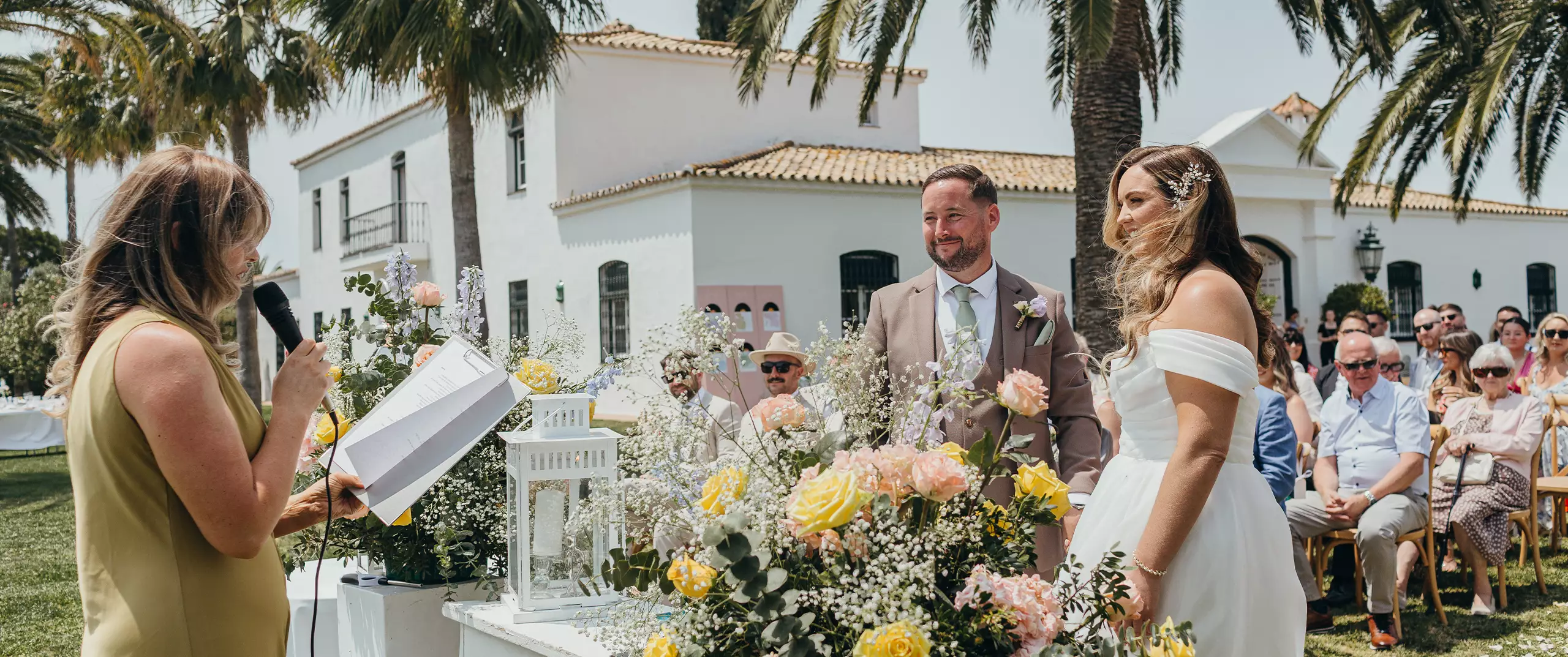 A wedding ceremony outdoors features a bride and groom standing before an officiant, with guests seated behind them and flowers arranged on the table in the foreground.