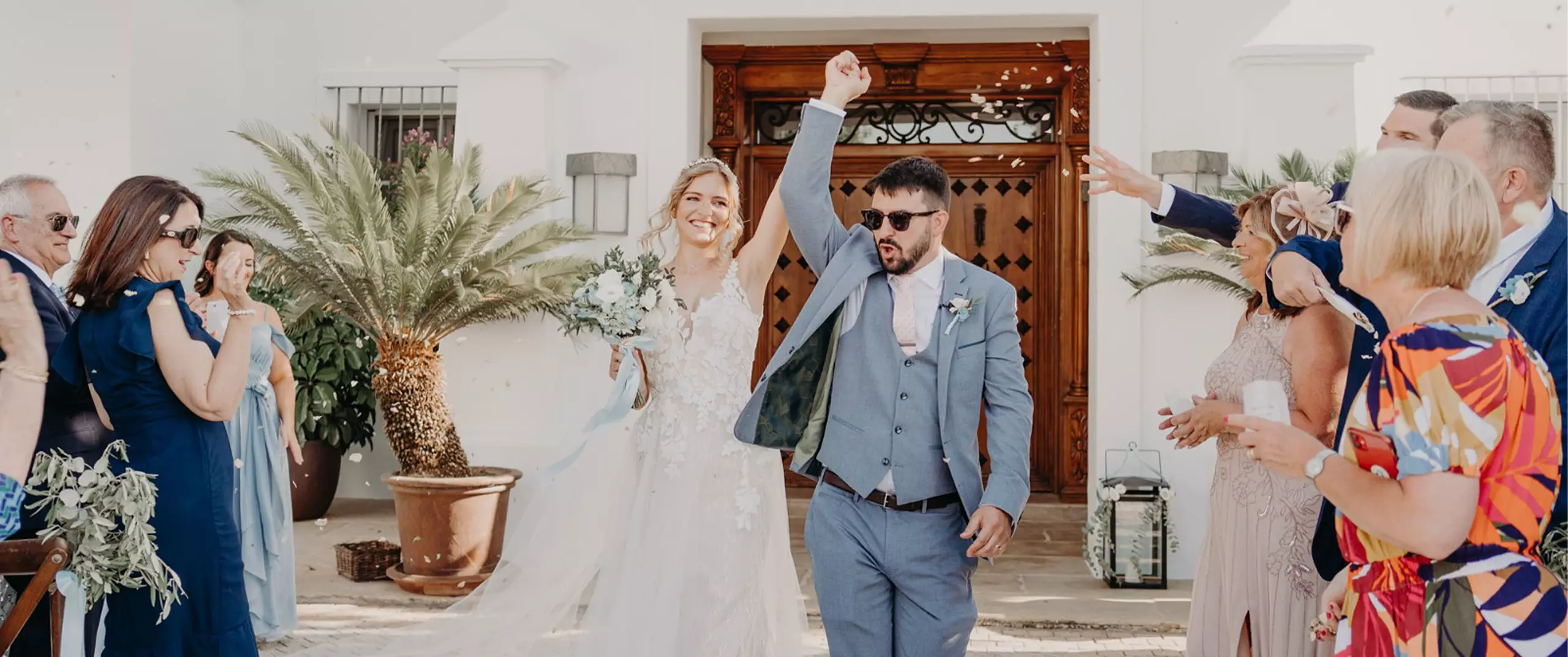 A bride and groom walk hand in hand outdoors, surrounded by guests who are cheering and tossing confetti.