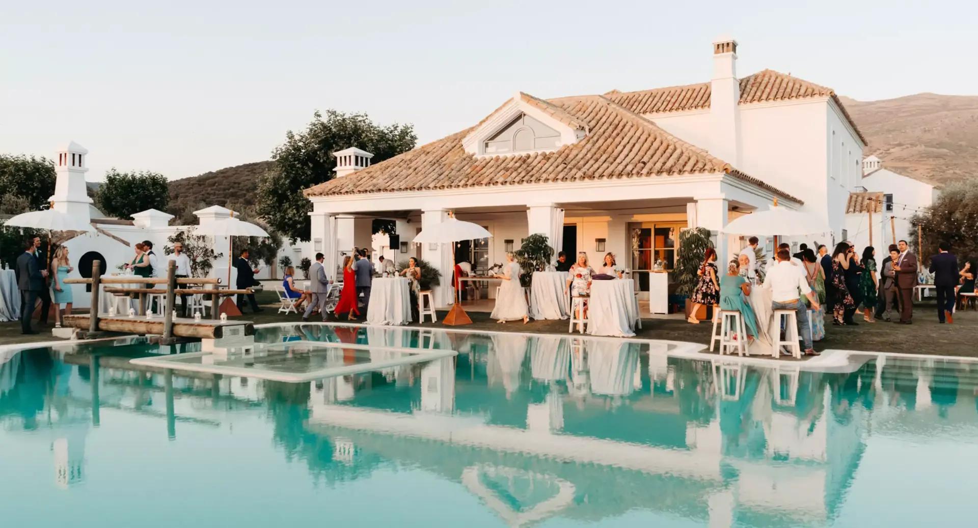 People gather around a swimming pool and outdoor seating area at a white villa during an event or celebration, with tables, umbrellas, and distant mountains visible.