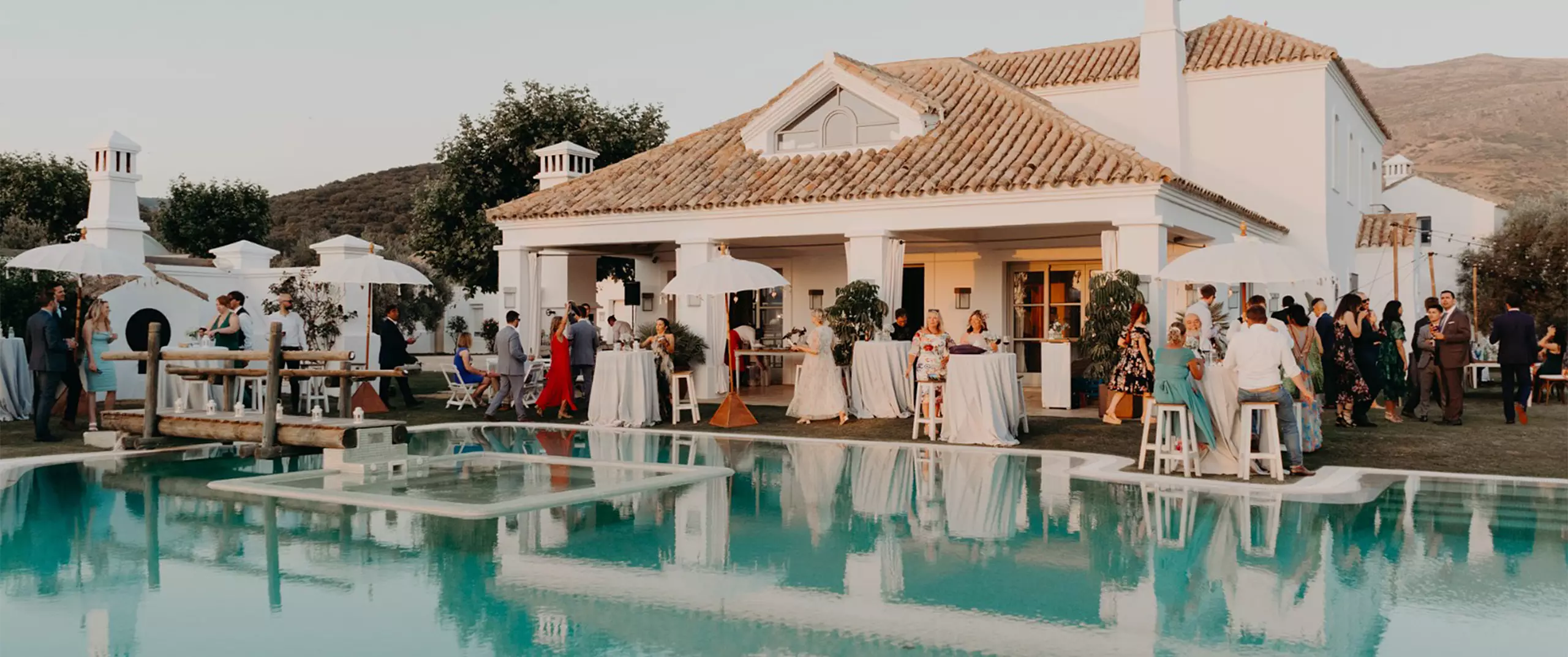 People gather around tables and stand near a swimming pool outside a large white house during a social event or party.