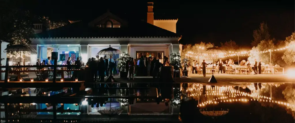 Nighttime outdoor event with people gathered near a lit building, string lights overhead, and reflections visible in a nearby pool.