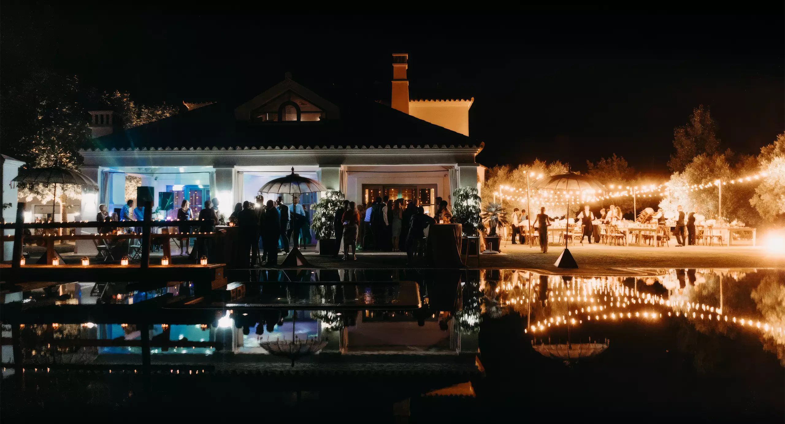 A nighttime outdoor event with guests gathered near a well-lit building, string lights, and reflections visible on a pool in the foreground.