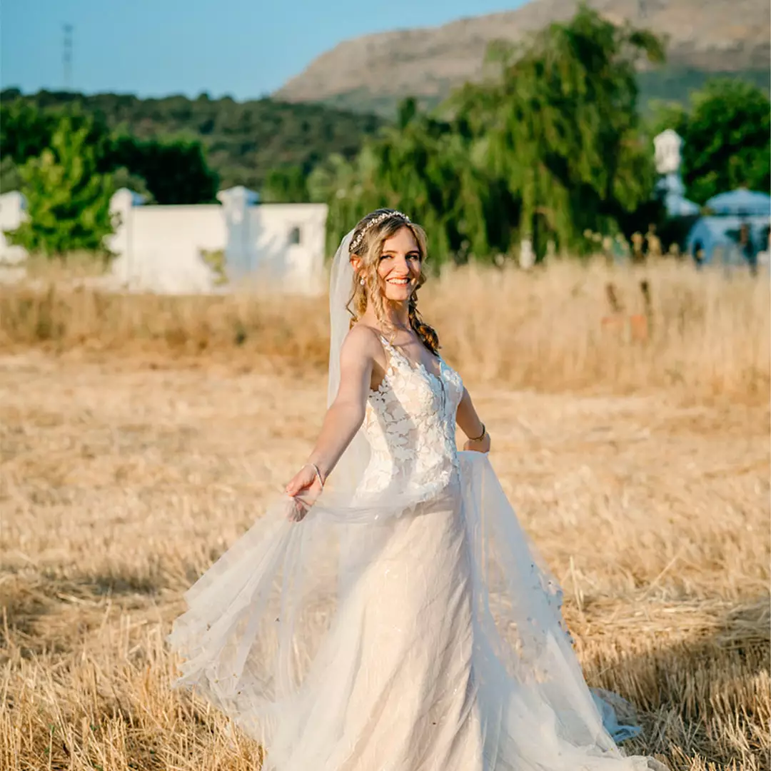A bride in a white wedding dress stands in a sunlit field, holding her veil and smiling, with trees and hills in the background.